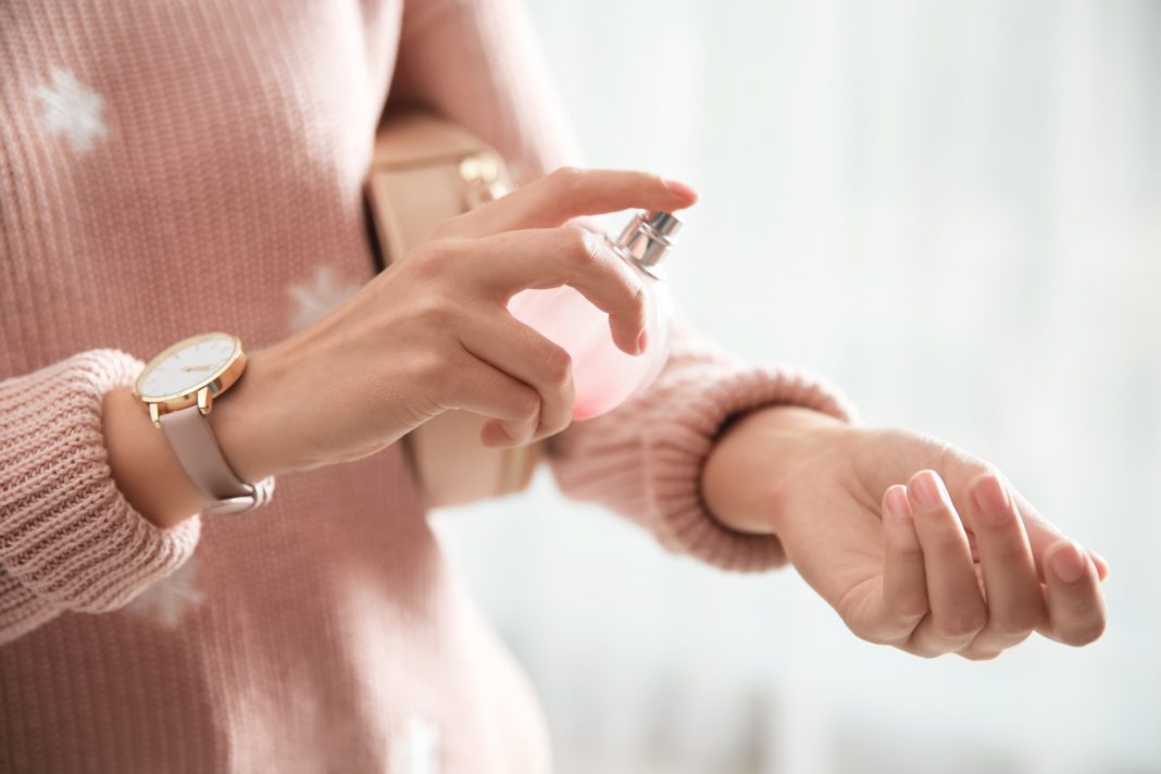 Eine Fru im rosa Pullover besprüht ihr Handgelenk mit einem Duft aus einem Zerstäuber, den sie in der anderen Hand hält.