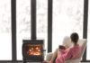Woman sitting with cup on chair by the fireplace at modern house on nature during winter time. Concept of winter mood and comfort at home. Girl wearing hat and pink sweater