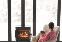 Woman sitting with cup on chair by the fireplace at modern house on nature during winter time. Concept of winter mood and comfort at home. Girl wearing hat and pink sweater