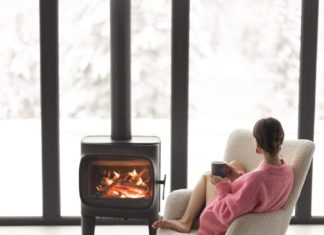 Woman sitting with cup on chair by the fireplace at modern house on nature during winter time. Concept of winter mood and comfort at home. Girl wearing hat and pink sweater