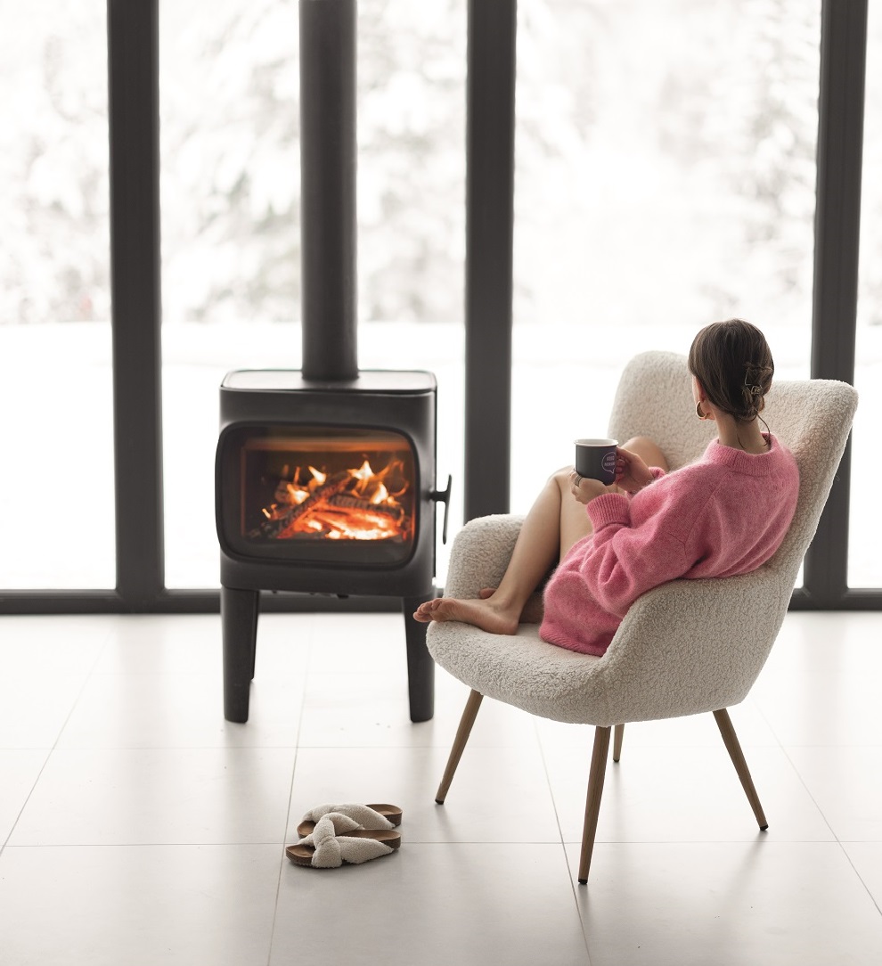 Woman sitting with cup on chair by the fireplace at modern house on nature during winter time. Concept of winter mood and comfort at home. Girl wearing hat and pink sweater