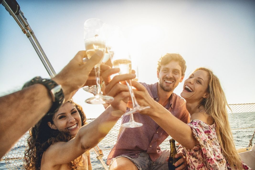 Zwei junge Frauen und Männer stoßen mit Sektgläsern an. Sie sind in einem Boot auf dem Wasser.