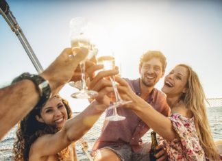 Zwei junge Frauen und Männer stoßen mit Sektgläsern an. Sie sind in einem Boot auf dem Wasser.