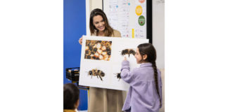 Angelina Jolie trägt eine Schautafel mit Bienenabbildungen in der Hand, die sie einem Mädchen erklärt.