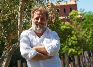 Frei sein und eins mit der Natur – Das Besondere an FKK-Camping Ein Mann im weißen Hemd lehnt sich mit verschränkten Armen gegen einen Holzzaun