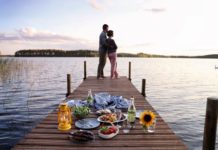 Eine Frau lehnt sich an einen Mann. Sie stehen auf einem Holzsteg über dem Wasser. Auf dem Holzsteg ist ein Picknick vorbereitet.