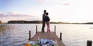 Eine Frau lehnt sich an einen Mann. Sie stehen auf einem Holzsteg über dem Wasser. Auf dem Holzsteg ist ein Picknick vorbereitet.