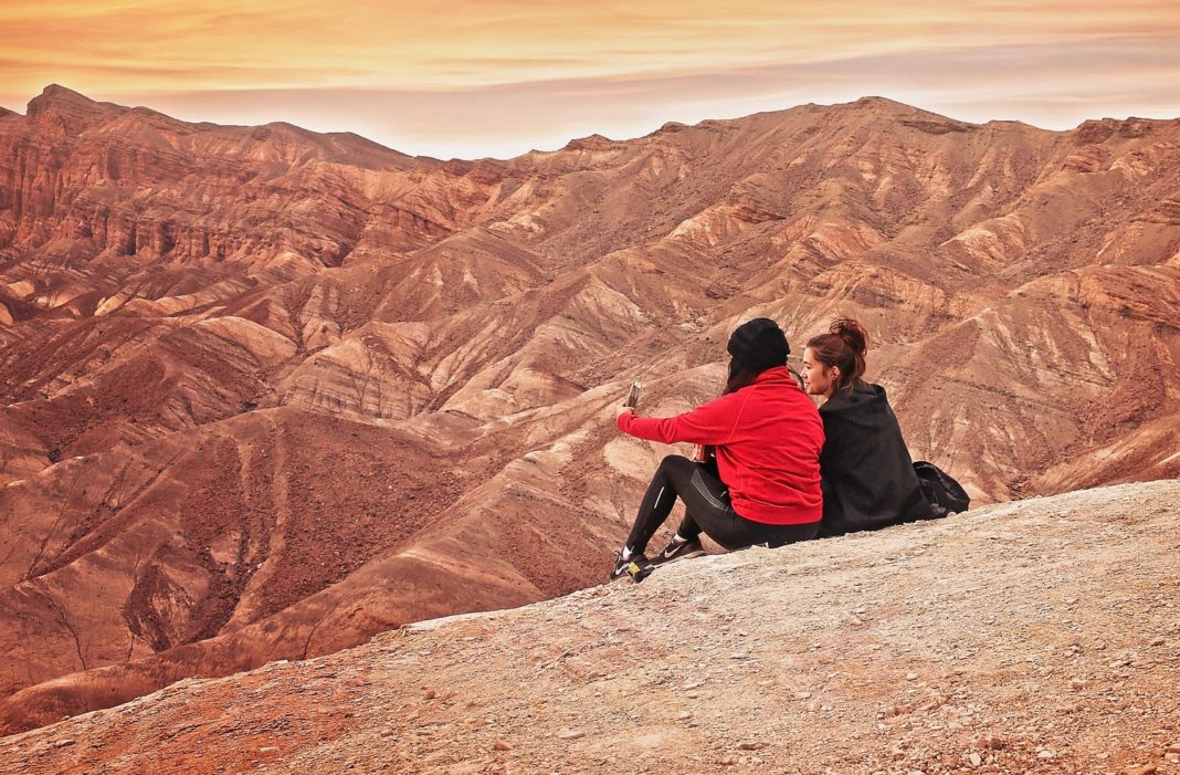 Zwei junge Frauen sitzen auf einer Felserhöhung in der Wüste und bewundern den Sonnenaufgang.