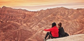 Zwei junge Frauen sitzen auf einer Felserhöhung in der Wüste und bewundern den Sonnenaufgang.