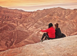 Zwei junge Frauen sitzen auf einer Felserhöhung in der Wüste und bewundern den Sonnenaufgang.