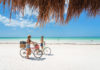 Isla Holbox, Yucatan, Mexico - April 9, 2021: Two adult females ride bikes on the white sand beach of the Gulf of Mexico on a beautiful day.