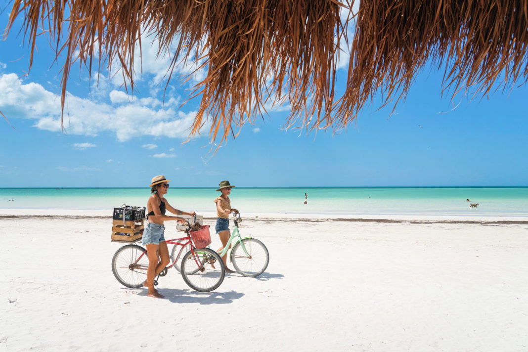 Isla Holbox, Yucatan, Mexico - April 9, 2021: Two adult females ride bikes on the white sand beach of the Gulf of Mexico on a beautiful day.