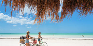 Isla Holbox, Yucatan, Mexico - April 9, 2021: Two adult females ride bikes on the white sand beach of the Gulf of Mexico on a beautiful day.