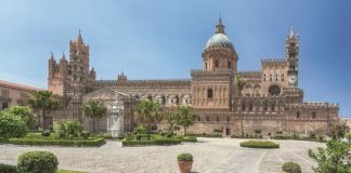 Bild von der Kathedrale von Palermo. Ein großer Bau mit blauer Kuppel. Davor ein gepflasterer Platz mit Blumenkübeln und Parkanlage.