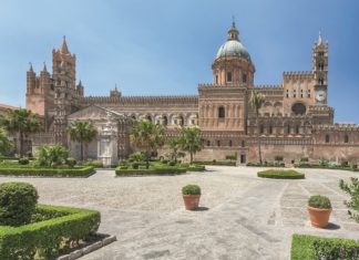 Bild von der Kathedrale von Palermo. Ein großer Bau mit blauer Kuppel. Davor ein gepflasterer Platz mit Blumenkübeln und Parkanlage.