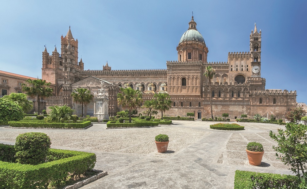 Bild von der Kathedrale von Palermo. Ein großer Bau mit blauer Kuppel. Davor ein gepflasterer Platz mit Blumenkübeln und Parkanlage.