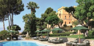 Blauen Pool mit Iegestühlen und Sonnenliegen vor einer mit Palmen bestandenen Villa.