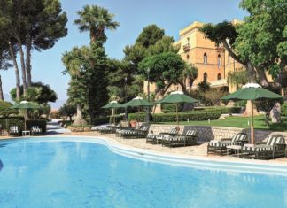Blauen Pool mit Iegestühlen und Sonnenliegen vor einer mit Palmen bestandenen Villa.