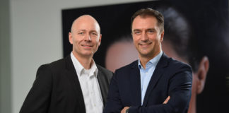 Zwei Männer in Business-Kleidung stehen in lockerer Pose vor einem Bild und blicken lächelnd in die Kamera.