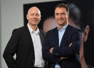 Zwei Männer in Business-Kleidung stehen in lockerer Pose vor einem Bild und blicken lächelnd in die Kamera.