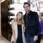 Eine Frau mit langen blonden Haaren und ein größerer Mann, beide in festlicher Kleidung, stehen vor einem Verkaufsregal und blicken in die Kamera.