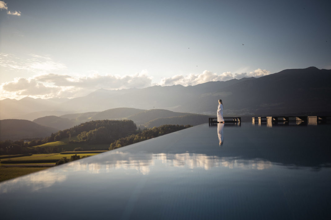 Eine Frau im weißen Bademantel steht am Rand eines Infinity-Pools und blickt auf ein Bergpanorama