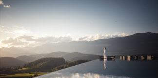 Eine Frau im weißen Bademantel steht am Rand eines Infinity-Pools und blickt auf ein Bergpanorama