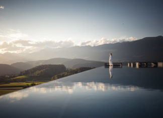 Eine Frau im weißen Bademantel steht am Rand eines Infinity-Pools und blickt auf ein Bergpanorama