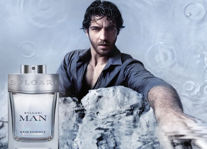 Ein dunkelhaariger Mann mit Bart greift über einen Felsen ins Wasser. Neben sich eine Flacon mit Parfum.