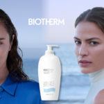 Zwei junge Frauen blicken in die Kamera. Zwischen ihnen eine Flasche Bodylotion, hinter ihnen das Meer.