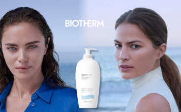 Zwei junge Frauen blicken in die Kamera. Zwischen ihnen eine Flasche Bodylotion, hinter ihnen das Meer.