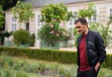 Ein Mann in dunkler Jacke und rotem Pullover steht in einem Garten mit Rosensträuchern im Hintergrund.