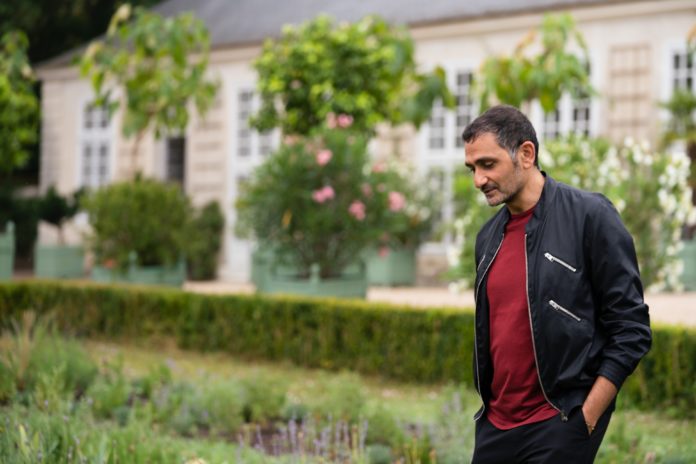 Ein Mann in dunkler Jacke und rotem Pullover steht in einem Garten mit Rosensträuchern im Hintergrund.