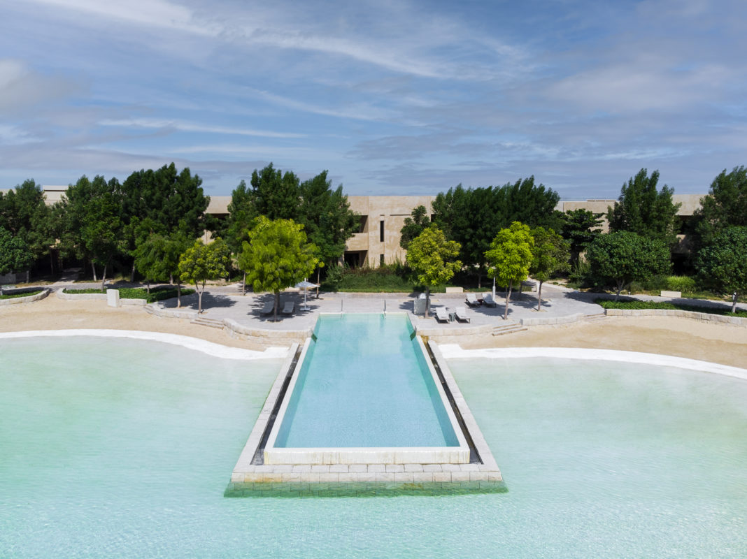 Blaues Meer, Strand, Bäume und das Hotel im Hintergrund. Ein langer eckiger Infinity-Pool ragt ins Meer. hinein.