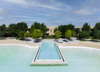 Blaues Meer, Strand, Bäume und das Hotel im Hintergrund. Ein langer eckiger Infinity-Pool ragt ins Meer. hinein.