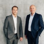 Zwei Männer im Anzug stehen nebeneinander vor einen betonfarbenen Hintergrund und blicken in die Kamera.