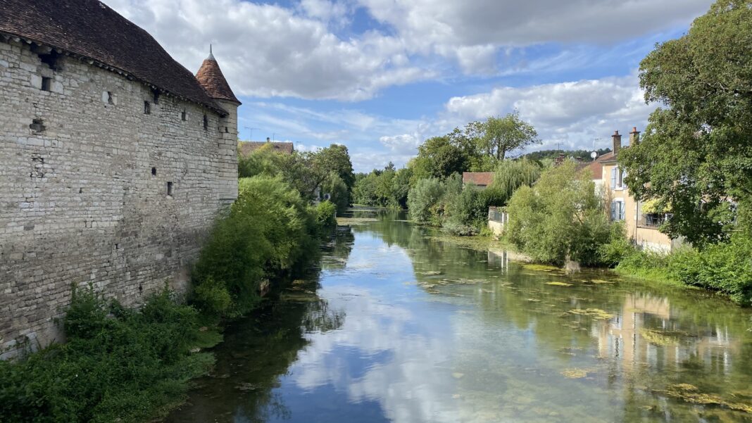 Blick auf einen Flußlauf, gesäumt von einer alten Mauer und Bewuchs. Der Himmel spiegelt sich im Wasser