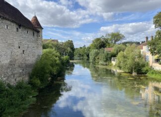 Blick auf einen Flußlauf, gesäumt von einer alten Mauer und Bewuchs. Der Himmel spiegelt sich im Wasser