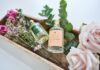 Zwei gläserne Parfumflakons in einem Holzbehältnis, umgeben von Eukalytuszweigen, Rosen und Zweigen mit kleinen lila Blüten.