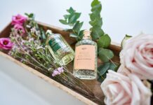 Zwei gläserne Parfumflakons in einem Holzbehältnis, umgeben von Eukalytuszweigen, Rosen und Zweigen mit kleinen lila Blüten.