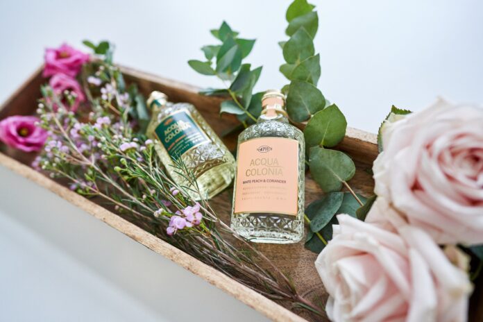 Zwei gläserne Parfumflakons in einem Holzbehältnis, umgeben von Eukalytuszweigen, Rosen und Zweigen mit kleinen lila Blüten.
