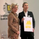 Zwei Frauen stehen vor einem Plakat. Eine der Frauen hält eine Urkunde in der Hand.