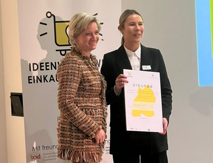 Zwei Frauen stehen vor einem Plakat. Eine der Frauen hält eine Urkunde in der Hand.