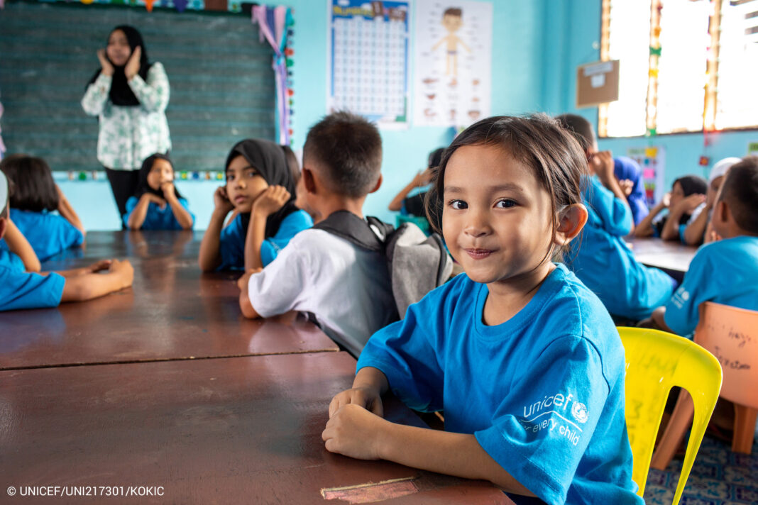 Ein kleines Mädchen in blauem T-Shirt sitzt in einem Klassenzimmer und schaut in die Kamera.