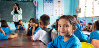 Ein kleines Mädchen in blauem T-Shirt sitzt in einem Klassenzimmer und schaut in die Kamera.