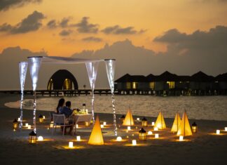 360 Grad Verwöhn-Programm – mit Gastkoch Eyck Zimmer im Januar im Outrigger Maldives Maafushivaru Resort Ein Paar am Strand bei Dämmerung, umgeben von stimmungsvoller Beleuchtung.