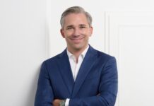 Neuer General Manager Lancôme DACH Ein weißhaariger, jüngerer Mann in einem blauen Anzug und weißem Hemd steht mit vor der Brust gekreuzten Armen vor einer weißen Tür.