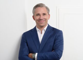 Neuer General Manager Lancôme DACH Ein weißhaariger, jüngerer Mann in einem blauen Anzug und weißem Hemd steht mit vor der Brust gekreuzten Armen vor einer weißen Tür.