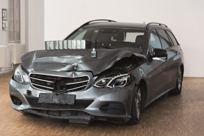 Graues Auto mit Frontschaden. Auf der zerbeulten Kühlerhaube ein Flacon des Parfums.