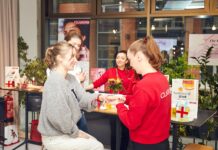 flaconi: Partnerschaft für die The Beauty Press Days Zwei junge Frauen sitzen an einem Messestand und lassen sich von zwei Frauen in roten Sweatshirts beraten.